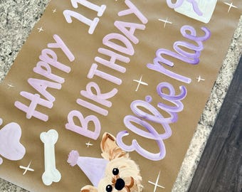 May include: A brown paper sign featuring hand-painted light purple text that reads Happy 11th Birthday Ellie Mae. The design includes illustrations of two balloons, a gift box, a dog bone, and a small dog wearing a light purple party hat.