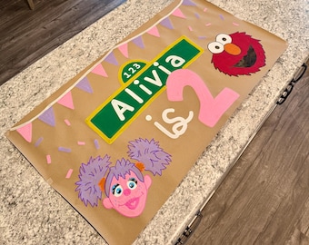 May include: A rectangular brown paper banner featuring a green street sign with the text 123 Alivia, a large pink number 2, and the text is in white. The banner includes illustrations of Elmo and Abby Cadabby, pink and purple bunting, and confetti.