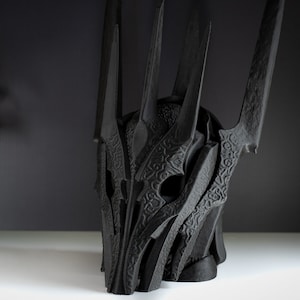 May include: A black, detailed, 3D printed helmet with a crown of spikes. The helmet is made of a textured material and has a detailed design.