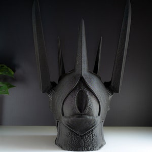 May include: A black, 3D printed crown with a pointed, angular design. The crown has a single, large eye-shaped opening in the front.