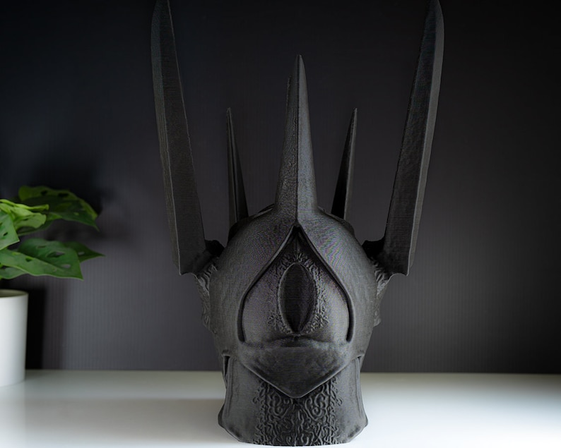 May include: A black, 3D printed crown with a pointed, angular design. The crown has a single, large eye-shaped opening in the front.