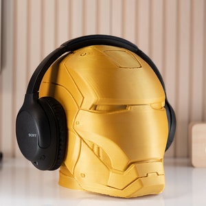 May include: A gold-colored Iron Man helmet-shaped headphone stand with black headphones resting on top. The stand is detailed with the helmet's design. A wooden clock and a tablet are in the background.