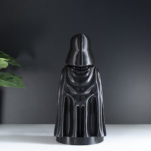 May include: A black plastic figurine of Darth Vader from Star Wars. The figurine is standing with its back to the camera and is wearing a black cape.