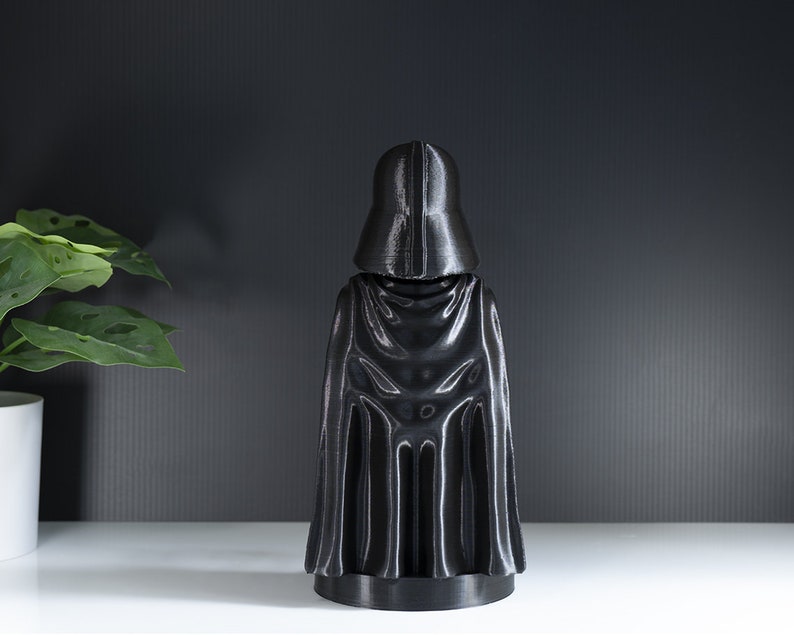May include: A black plastic figurine of Darth Vader from Star Wars. The figurine is standing with its back to the camera and is wearing a black cape.