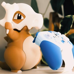 Może przedstawiać: Figurkę Pokemon w kolorze brązowym i białym z niebieskim i białym kontrolerem do gier wideo.