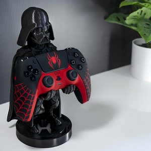 May include: A black Darth Vader figurine holding a red and black PlayStation 5 controller with a spider web design. The controller is on a black stand.
