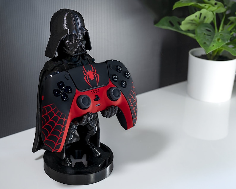 May include: A black Darth Vader figurine holding a red and black PlayStation 5 controller with a spider web design. The controller is on a black stand.