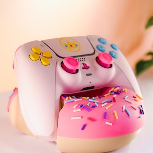 Pode incluir: Um controle de videogame rosa claro com botões amarelos, azuis e rosa. O controle é decorado com uma coroa amarela e é combinado com um donut rosa com granulado colorido.