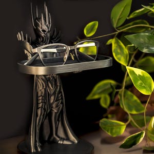 May include: A black and silver 3D printed stand shaped like a fantasy figure holding a pair of glasses on a tray. The stand is on a dark surface with a green plant in the background.