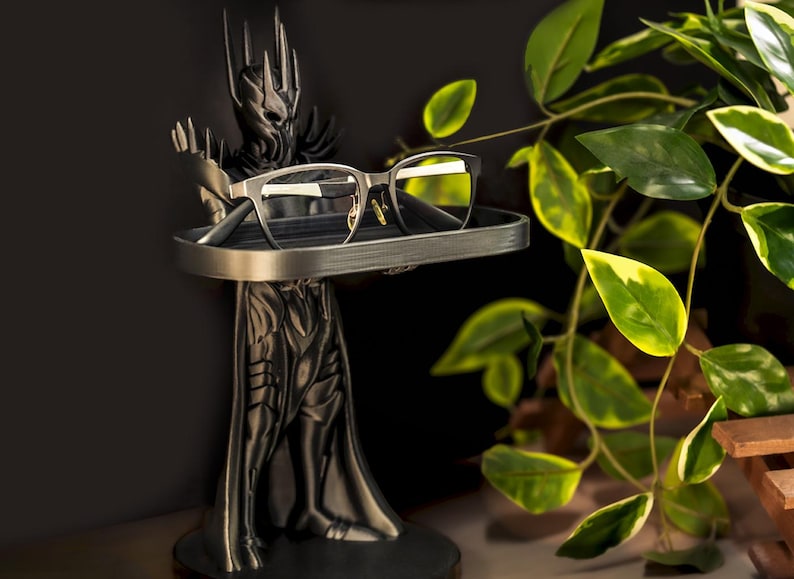 May include: A black and silver 3D printed stand shaped like a fantasy figure holding a pair of glasses on a tray. The stand is on a dark surface with a green plant in the background.