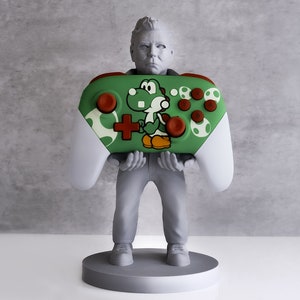Könnte beinhalten: Eine graue Figur eines Menschen, der einen grün-weißen Nintendo Switch-Controller mit einem Yoshi-Charakterdesign hält. Der Controller hat braune Knöpfe und eine weiße Basis.