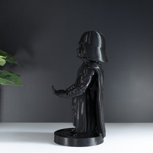 May include: A black plastic Darth Vader phone stand. The stand is designed to hold a smartphone in a vertical position.