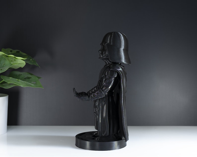 May include: A black plastic Darth Vader phone stand. The stand is designed to hold a smartphone in a vertical position.