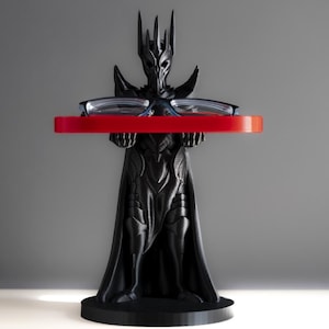 May include: A black 3D printed figurine of a fantasy character wearing a crown and holding a red tray with a pair of glasses on top. The figurine is standing on a black base.