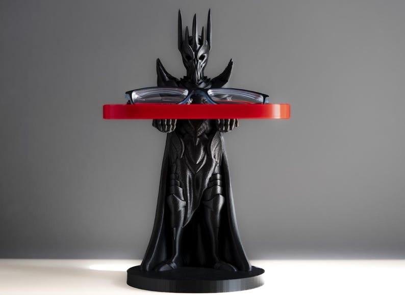 May include: A black 3D printed figurine of a fantasy character wearing a crown and holding a red tray with a pair of glasses on top. The figurine is standing on a black base.