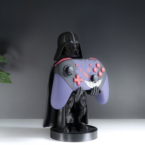 May include: A black Darth Vader figurine holding a purple and black video game controller with red buttons. The controller has a white and red smiley face design.