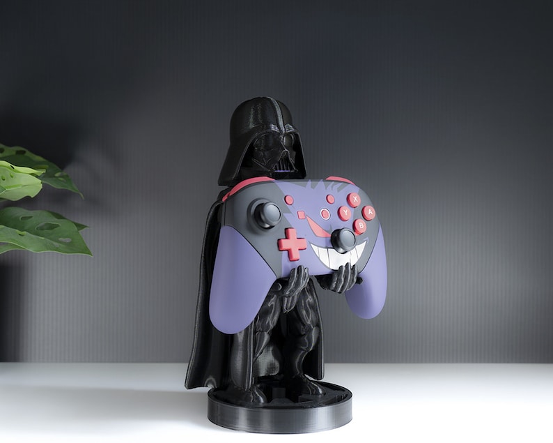 May include: A black Darth Vader figurine holding a purple and black video game controller with red buttons. The controller has a white and red smiley face design.