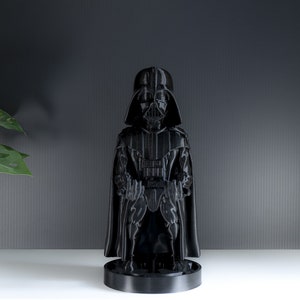 May include: A black Darth Vader figurine with a cape and a black base. The figurine is standing on a white surface.
