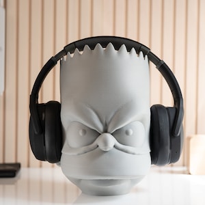 May include: A gray, 3D-printed headphone stand shaped like a cartoon character's head, with a black pair of headphones resting on it. The stand has an angry expression and a jagged edge at the top. The headphones are black.