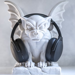 May include: A grey stone gargoyle sculpture wearing black over-ear headphones. The gargoyle has wings and a menacing expression.