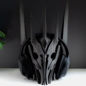 May include: A black 3D printed headphone stand shaped like a crown. The stand has intricate details and is designed to hold over-ear headphones.