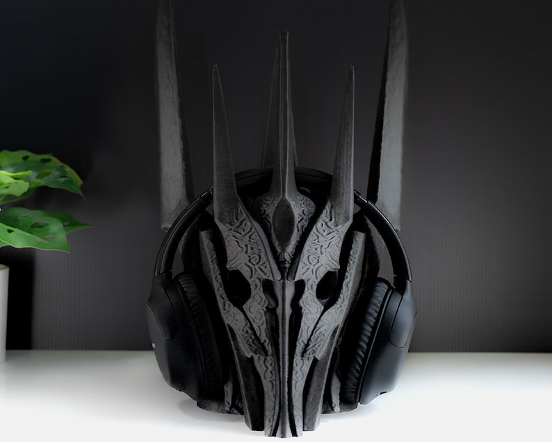 May include: A black 3D printed headphone stand shaped like a crown. The stand has intricate details and is designed to hold over-ear headphones.