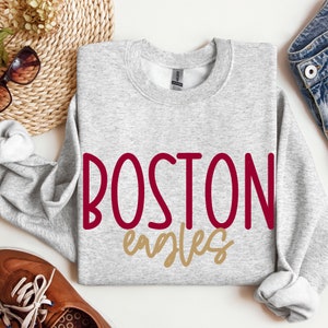 May include: A light gray sweatshirt with the words "BOSTON EAGLES" in burgundy and gold lettering. The sweatshirt is laid flat on a white surface, with a pair of sunglasses, a woven basket, and a pair of brown shoes in the background.