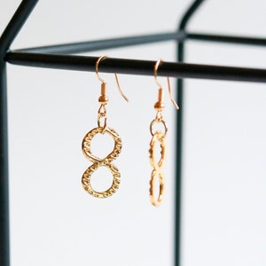 Puede incluir: Un par de pendientes de oro con forma de símbolo de infinito colgados de un expositor de metal negro. Los pendientes son texturizados y tienen un diseño delicado.