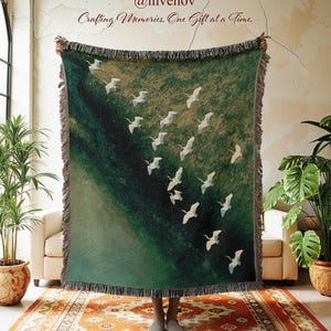May include: A woven throw blanket featuring a dark green landscape with a flock of white birds in flight. The blanket has a fringed edge and displays the text @invenov and Crafting Memories, One Gift at a Time at the top center.
