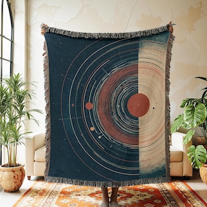 May include: A navy blue woven throw blanket with a celestial design. The blanket features a circular pattern in rust, beige, and white. It has a fringed edge. The blanket is held up in front of a person.
