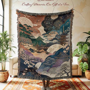 May include: A woven throw blanket featuring a detailed Japanese-inspired landscape with a pagoda, a crane, a bridge, and floral motifs in shades of blue, green, and brown. The blanket has a fringed edge and includes the text @invenov Crafting Memories, One Gift at a Time.