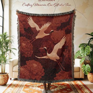 May include: A burgundy throw blanket featuring a crane design with white wings and red accents. The blanket has a floral pattern and a fringed edge. The text "@invenov Crafting Memories, One Gift at a Time." is visible.