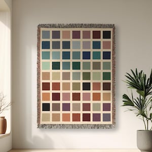May include: A woven throw blanket with a grid pattern of squares in various colours, including blue, green, brown, and purple. The blanket is displayed on a white wall in a room with a large window and a potted plant.