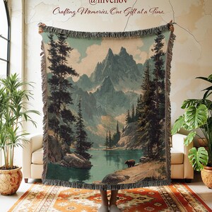 May include: A woven tapestry blanket featuring a scenic landscape with mountains, a lake, and trees. The blanket has a fringed edge and is held up in front of a person. The text "@invenov Crafting Memories, One Gift at a Time." is visible.