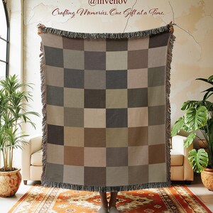 May include: A woven blanket with a checkerboard pattern in shades of brown, gray, and beige. The blanket has a fringed edge and is held up in front of a window. The text "@invenov Crafting Memories, One Gift at a Time." is visible.