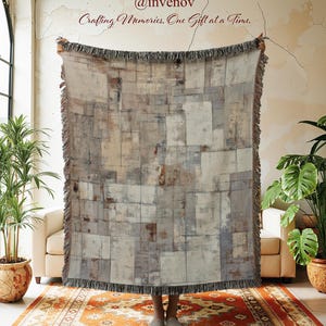 May include: A rectangular woven throw blanket with a geometric pattern in shades of gray, beige, and brown. The blanket has a fringed edge and is held up by a person. The text "@invenov Crafting Memories, One Gift at a Time." is visible.