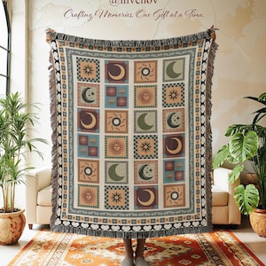 May include: A woven throw blanket featuring a repeating pattern of suns, moons, and stars in squares. The blanket has a cream background with brown, green, and blue accents. The blanket has a fringed edge and the text "Crafting Memories, One Gift at a Time."