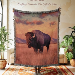 May include: A woven throw blanket featuring a large bison standing in a grassy field under a sunset sky. The blanket has fringed edges and displays the text @invenov and Crafting Memories, One Gift at a Time at the top center.