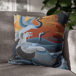 May include: A square throw cushion featuring a printed design of two white cranes with blue and yellow wings flying over stylised dark blue waves and a deep orange background with a large golden sun. The fabric has a textured appearance.