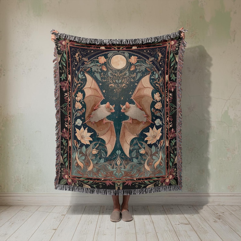 May include: A woven tapestry blanket featuring a symmetrical design of two bats with outstretched wings, set against a teal background with floral and celestial elements. The blanket has a fringed edge and a full moon.