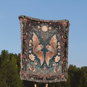 May include: A dark teal throw blanket with a symmetrical design featuring two bats with outstretched wings, a full moon, and floral accents. The blanket has a fringed edge and is held up against a blue sky and green trees.