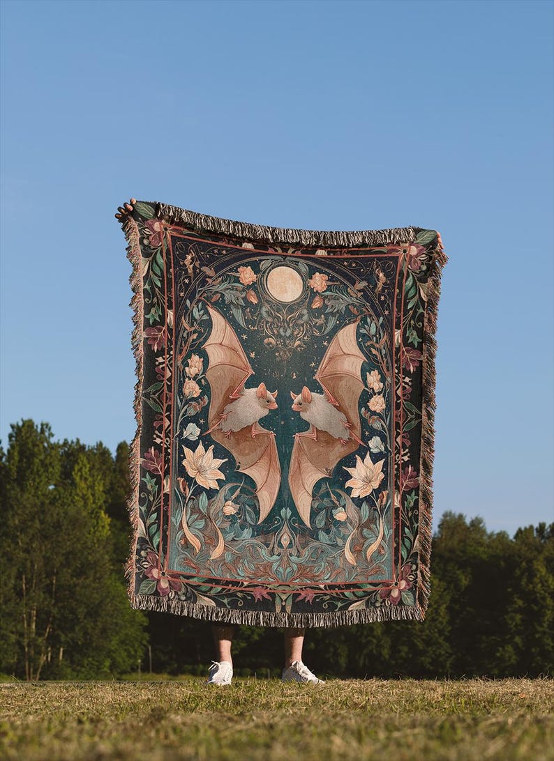 May include: A dark teal throw blanket with a symmetrical design featuring two bats with outstretched wings, a full moon, and floral accents. The blanket has a fringed edge and is held up against a blue sky and green trees.