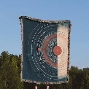 May include: A rectangular throw blanket with a celestial design. The blanket is split vertically, with a dark teal side featuring a planetary diagram and a beige side. The blanket has a fringed edge and is held up against a blue sky.