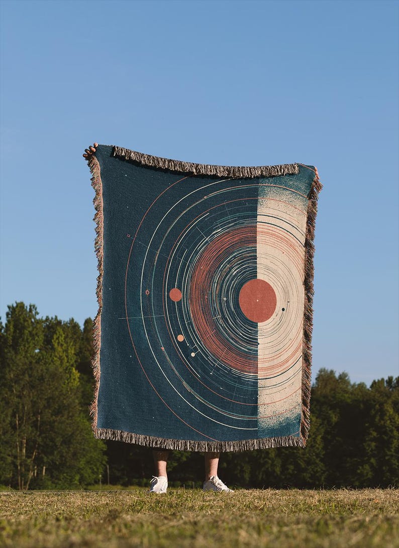 May include: A rectangular throw blanket with a celestial design. The blanket is split vertically, with a dark teal side featuring a planetary diagram and a beige side. The blanket has a fringed edge and is held up against a blue sky.