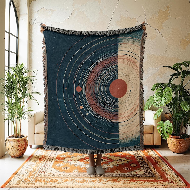 May include: A navy blue woven throw blanket with a celestial design. The blanket features a circular pattern in rust, beige, and white. It has a fringed edge. The blanket is held up in front of a person.