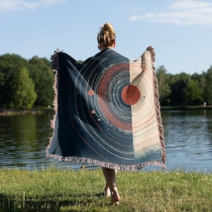 May include: A woven blanket with a celestial design, featuring concentric circles in shades of navy, rust, and beige. The blanket has a fringed edge and is held up against a backdrop of a lake and trees.