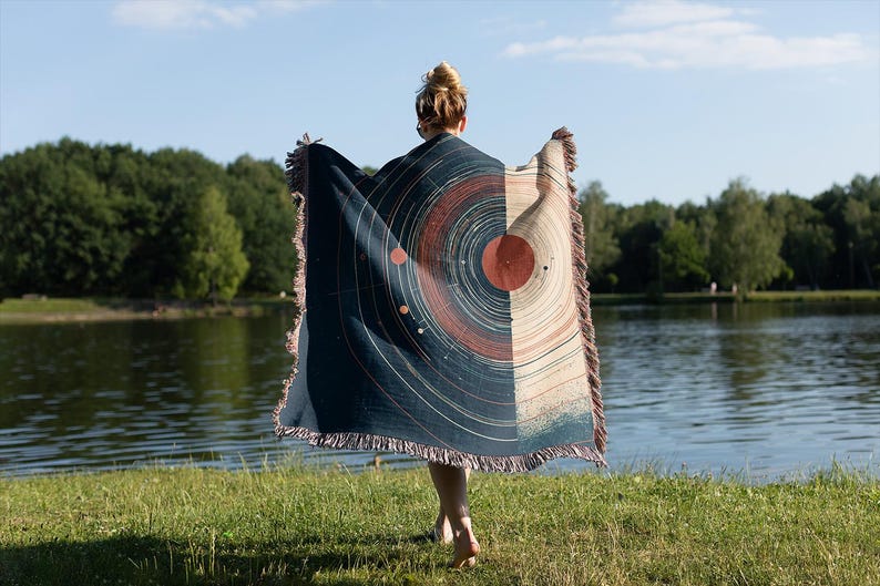 May include: A woven blanket with a celestial design, featuring concentric circles in shades of navy, rust, and beige. The blanket has a fringed edge and is held up against a backdrop of a lake and trees.