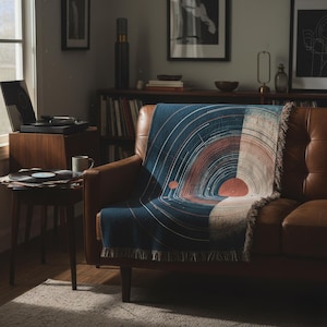May include: A navy blue throw blanket with a circular design in shades of orange, white, and blue is draped over a brown leather sofa. The blanket has a fringed edge. A record player and records are visible in the background.