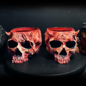 May include: Two red and flesh-toned skull-shaped planters with horns, set on a black surface. The planters have large eye sockets and a textured, weathered appearance. A black skull is visible to the left, and a jar with a skull on top is to the right.