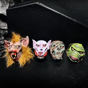 May include: Four small monster head figurines arranged in a row. The first is a furry creature with yellow hair and sharp teeth, the second is a pale vampire with a bat pendant, the third is a wrapped mummy, and the fourth is a green creature.